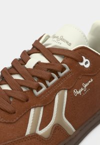 Καφέ δερμάτινο αθλητικό παπούτσι με εκρού λεπτομέρειες, υφασμένα κορδόνια και λογότυπο. Διαθέτει αθλητικό σχέδιο με διακριτικό branding και ραμμένα άκρα.