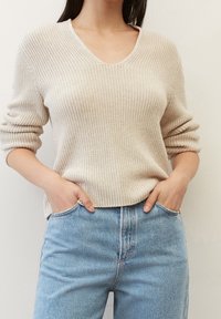 Beżowy sweter dziewiarski z dekoltem w serek, z długimi rękawami i luźnym krojem, zestawiony z jasnoniebieskimi jeansami o wysokim stanie. Gładka faktura, minimalistyczny design.