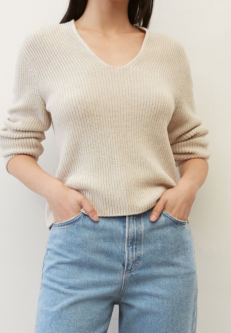 Beżowy sweter dziewiarski z dekoltem w serek, z długimi rękawami i luźnym krojem, zestawiony z jasnoniebieskimi jeansami o wysokim stanie. Gładka faktura, minimalistyczny design.