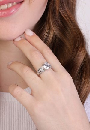 Bague de fiançailles en diamant avec une grande pierre centrale, un anneau en argent et des accents détaillés. Mains douces sur un fond clair.
