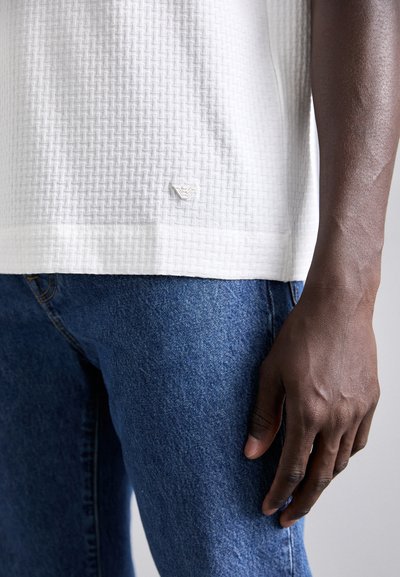 Chemise blanche à texture avec un motif tissé, dotée d'une petite étiquette logo à l'ourlet, associée à un jean en denim bleu.