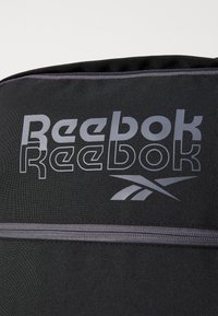 Reebok sporta soma melnā audumā ar sudraba logotipu un rāvējslēdzēju. Vienmērīga tekstūra ar horizontālām svītrām pa somas virsmu.