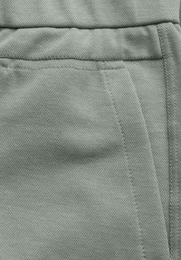 Jasnozielona tkanina z wyraźnym wzorem o gładkim wykończeniu. Widoczne szwy i szczegóły przeszyć na krawędziach, wskazujące na konstrukcję odzieży.