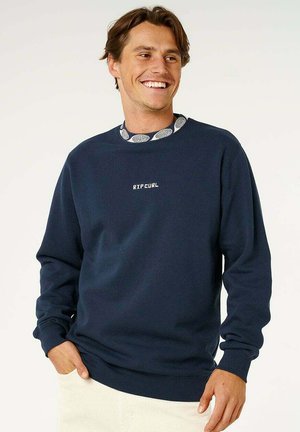 Uśmiechnięty mężczyzna w granatowej bluzie Rip Curl na wzorzystej koszuli, z jedną ręką w kieszeni kremowych spodni, na jednolitym tle.