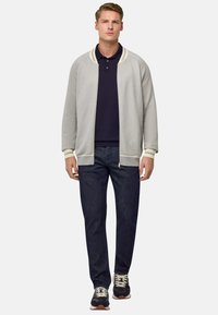 Giacca zip-up grigia chiara con accenti crema, abbinata a una polo blu navy, jeans scuri e sneakers nere con lacci bianchi e suole marroni.