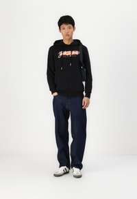 Sudadera negra con logo, cordones, puños acanalados, combinada con jeans sueltos de color azul oscuro y zapatillas blancas con franjas negras.