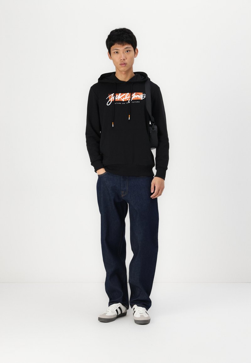Sudadera negra con logo, cordones, puños acanalados, combinada con jeans sueltos de color azul oscuro y zapatillas blancas con franjas negras.