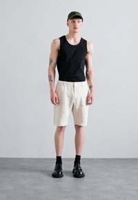 Schwarzes Tanktop, hellbeige Shorts, schwarze Schuhe und eine grüne Cap. Glatter Stoff, entspannter Schnitt und sichtbare Tattoos an den Armen.