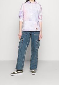 Hoodie ma pastelowy wzór tie-dye z przednią kieszenią; zestawiony z luźnymi niebieskimi spodniami cargo i zielonymi trampkami z białymi akcentami.