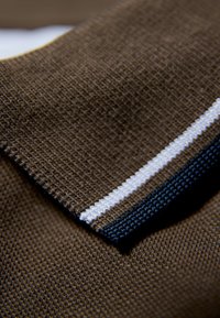 Brun pikétröja i tyg med texturerad yta, med en krage som har vita och mörkblå ränder. Närbild visar söm detaljer.