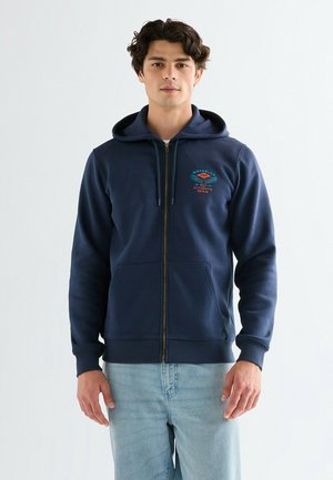 Młody mężczyzna w granatowej bluzie z kapturem na suwak, z przednimi kieszeniami i logo, w jasnoniebieskich dżinsach, stojący na jednolitym tle.