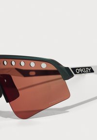 Grüne und weiße Sonnenbrille mit großen rot getönten Gläsern, die über kreisförmige Ausschnitte und das "Oakley"-Logo im Gestell verfügt.