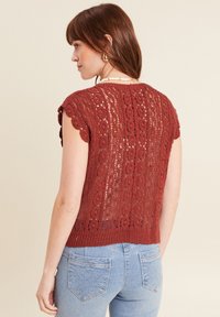 Top corto a maniche corte in pizzo rosso con bordi a scallop. Design a rete e motivo testurizzato. Indossato con jeans blu chiaro con tasche decorative.
