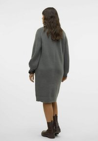 Vestido de suéter gris de punto con mangas largas texturizadas, aberturas laterales y dobladillo acanalado. La modelo lleva botas de tobillo oscuras y gruesas.