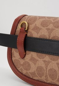 Brun mønstret stof taske med sort læderrem og røde læderdetaljer. Har guld hardware og kontraststing detaljer.