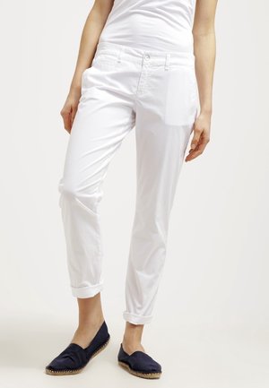 Vrouw draagt witte opgerolde broek, wit topje en marineblauwe espadrilles, staand tegen een effen lichte achtergrond.