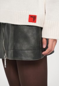 Maglione bianco a maglia con etichetta rossa Max&Co, indossato sopra una gonna di pelle nera con zip frontale, abbinato a collant trasparenti marroni scuri e con una mano appoggiata sulla gonna.