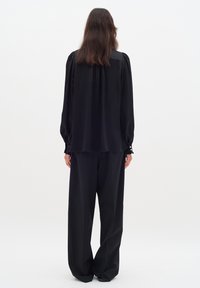 Musta pitkähiha blouse, jossa on rypytetyt hartiasaumet, löysä istuvuus ja napitettu hihansuu, yhdistettynä leveisiin mustiin housuihin, sileä pinta.