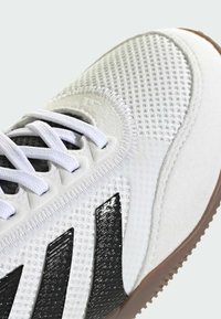 Close-up van een witte sport schoen met mesh stof, witte veters, zwarte streepdétails en een getextureerde gumzool op een lichte achtergrond.