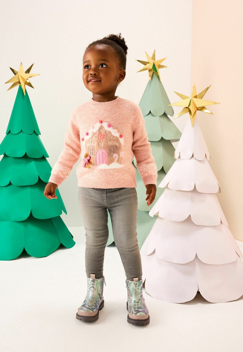 Barn iført pink julesweater og grå bukser står ved festlige papirltræer med guldstjerner på toppen.
