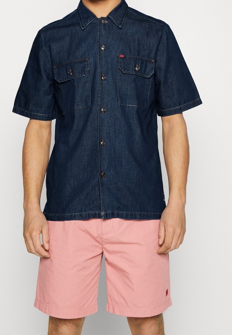 Homme portant une chemise en denim bleu foncé à manches courtes avec des poches à boutons et un short rose clair jusqu'aux genoux.