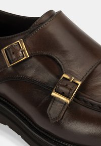Braune Lederschuhe mit zwei goldenen Schnallen, genähten Details entlang der Kanten und einem runden Zehendesign mit einer strukturierten Oberfläche.