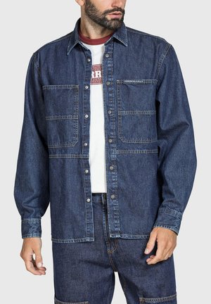 Uomo che indossa una giacca in denim blu scuro con grandi tasche frontali, camicia bianca con stampa rossa e pantaloni in denim blu scuro abbinati.