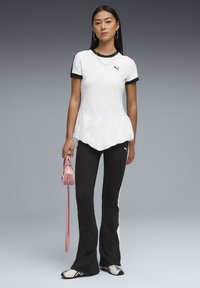 Γυναίκα με λευκό πουκάμισο με βολάν και κοντό μανίκι, μαύρο παντελόνι καμπάνα με λευκή ρίγα, αθλητικά παπούτσια, κρατώντας μικρή ροζ τσάντα, μπροστά σε γκρι φόντο.