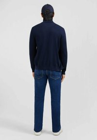 Pull en maille marine avec un col haut, associé à un jean en denim bleu. La tenue comprend des baskets blanches et une casquette bleue. Vue de dos affichée.