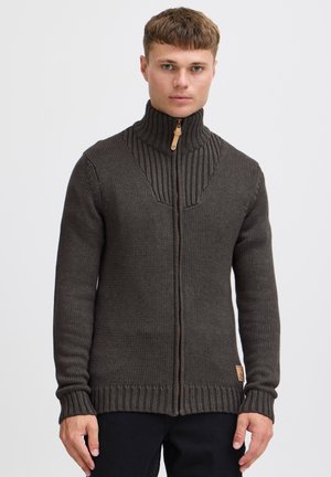 Grosse agrafe marron foncé avec col haut, design côtelé sur les épaules et tricot texturé. Présente une étiquette en cuir sur le bas.
