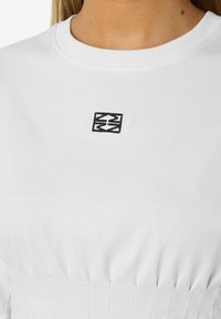 Balta marškinėliai su apvaliu kaklu, turintys mažą juodą geometrinį logotipą ant krūtinės ir susiraukšlėjusią tekstūrą apačioje.