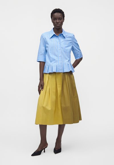 Femme portant une chemise bleue à manches courtes avec un ourlet plissé et une jupe midi plissée jaune, debout devant un fond blanc uni.