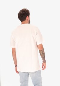 Homme avec un bras droit tatoué et des boucles d'oreilles, portant un t-shirt blanc uni et un jean bleu clair, se tenant de dos sur un fond blanc.