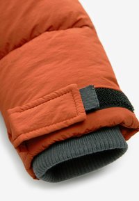 Manche de veste matelassée couleur orange brûlé avec un poignet côtelé gris et une sangle en Velcro noire. Texture lisse avec une apparence légère.