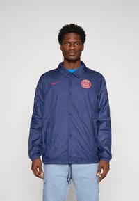 Man wearing a navy blue Paris Saint-Germain jacket with red logo and light gray sweatpants, standing against a plain background.
