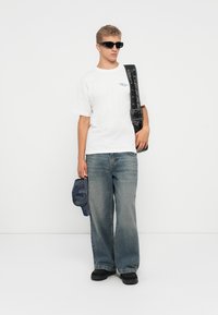 Hvit t-skjorte med blå logo, vide blå jeans, svarte solbriller, svart skulderveske og svarte sko. Hvit bakgrunn.