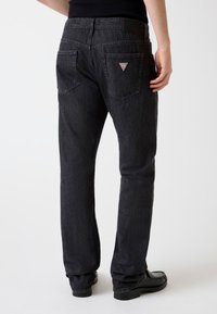 Jean en denim foncé avec une coupe droite, présentant une texture subtile, deux poches arrière et une petite écusson triangulaire au-dessus de la poche droite.