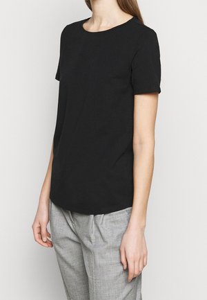 Femme portant un t-shirt noir uni à manches courtes et un pantalon gris clair, se tenant devant un fond clair uni.