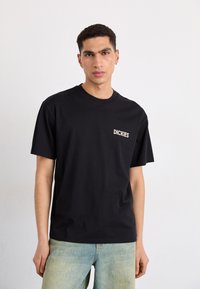 Camiseta de algodón negra de manga corta y cuello redondo. Cuenta con un pequeño logo blanco en el lado izquierdo del pecho. Combinada con un vaquero azul claro.