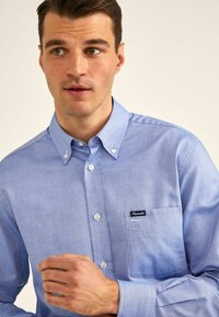 Camisa de botón azul claro hecha de una tela suave. Presenta un bolsillo en el pecho con una etiqueta, cuello puntiagudo y botones blancos.