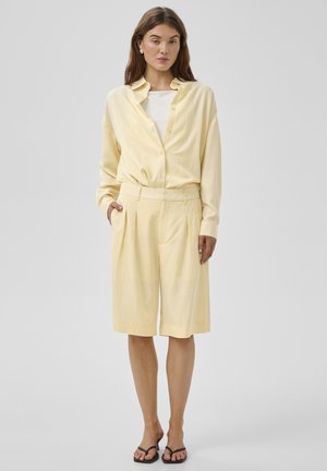 Nő világos sárga gombos ingben, hozzáillő térdig érő rövidnadrágban, fehér alsó felsőben és fekete magassarkú szandálban, szürke háttér előtt állva.