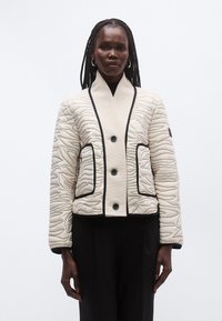 Sieviete ar ieveidotiem matiem, ģērbta krēmkrāsas pūderētu jaku ar melnu apmali, lielām pogām un sānu kabatām, valkā melnas bikses.