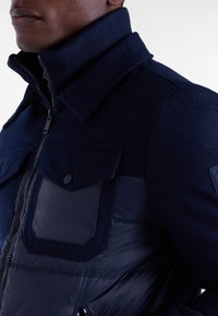 Marynarka w kolorze granatowym, wykonana z połączenia teksturowanej wełny i błyszczącego nylonu, z wysokim kołnierzem, zapięciem na zamek błyskawiczny oraz kieszeniami z klapami na klatce piersiowej.