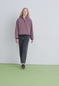 Chaqueta de forro polar con cremallera en color malva, con un cuello alto, cierre de cremallera y puños negros; combinada con jeans oscuros holgados y zapatos negros.