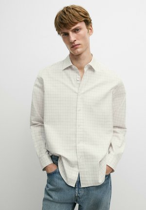 Ung mand med kort lyst brunt hår iført en hvid langærmet skjorte med et subtilt ternet mønster og blå jeans.