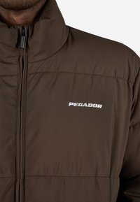 Brun polstret jakke med høy krage, med glidelås foran og "PEGA DOR"-logoen i hvitt på venstre side.