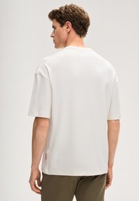 Biały T-shirt z krótkim rękawem o luźnym kroju, z okrągłym dekoltem i małym czerwonym logo na boku. Gładka bawełniana tekstura.