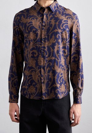 Man draagt een donkerblauw overhemd met knopen met bruine ingewikkelde bloemen- en wijnrankenpatronen, gecombineerd met een zwarte broek tegen een effen achtergrond.