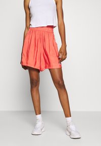 Korallfärgade satinshorts med ett elastiskt midjeband, featuring sidostråkar och en avslappnad passform. Bärs med en kortärmad vit linne och vita sneakers.