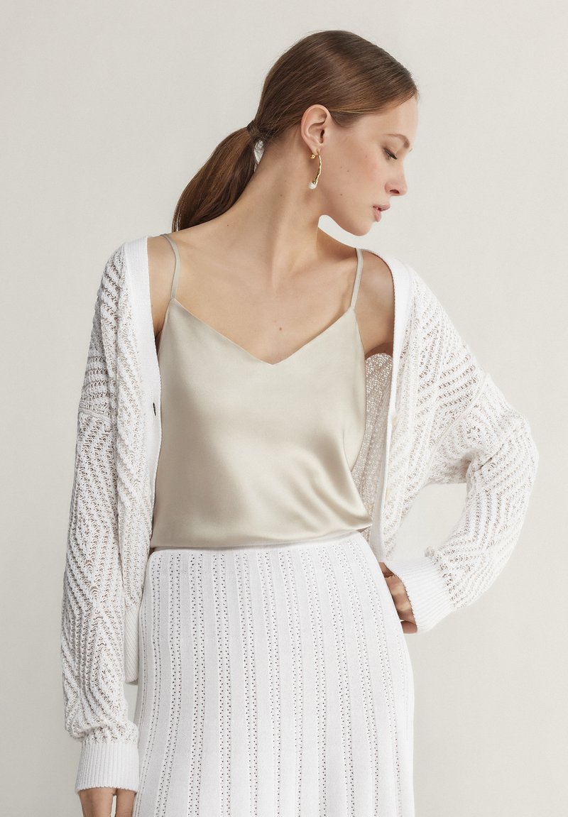 Femme aux cheveux bruns attachés en queue de cheval, portant une boucle d'oreille créole en or, un caraco en satin beige, un cardigan en maille blanc et une jupe blanche à motifs.